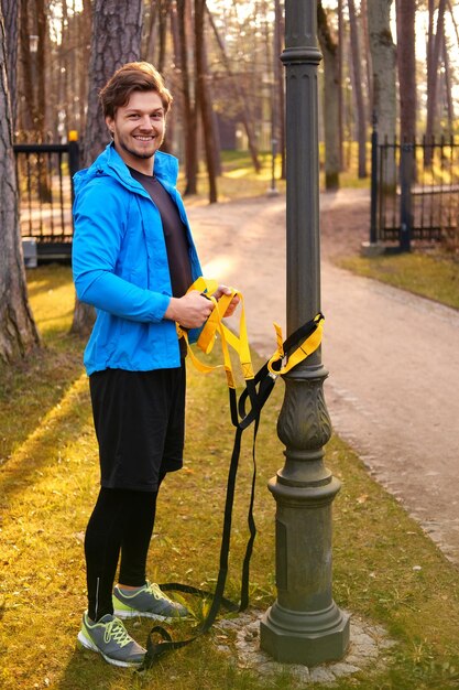 Modelo de fitness de homem em uma jaqueta esporte azul fazendo exercícios com tiras de fitness trx em um parque.
