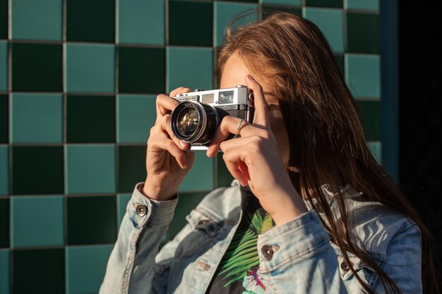 Modelo de garota legal com câmera de filme retrô vestindo uma jaqueta jeans, cabelo escuro ao ar livre sobre a muralha da cidade em um fundo de gaiola. Se divertindo na cidade com câmera, foto de viagem do fotógrafo.