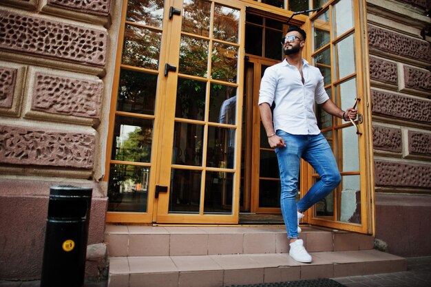 Modelo de homem árabe alto elegante em jeans de camisa branca e óculos de sol posou na rua da cidade Barba atraente cara árabe contra a porta de entrada do restaurante
