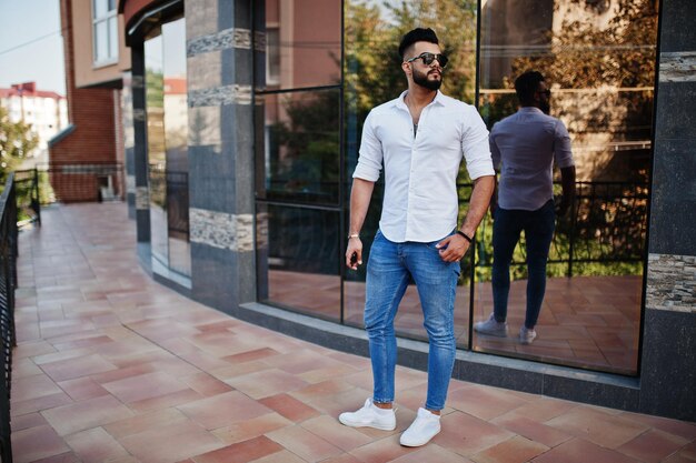 Modelo de homem árabe alto elegante em jeans de camisa branca e óculos de sol posou na rua da cidade Barba atraente cara árabe contra o edifício moderno