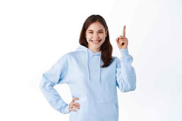 Modelo de jovem atraente em roupa casual, apontando o dedo de lado, mostrando seu texto promocional no espaço da cópia superior, sorrindo para a câmera em pé contra um fundo branco