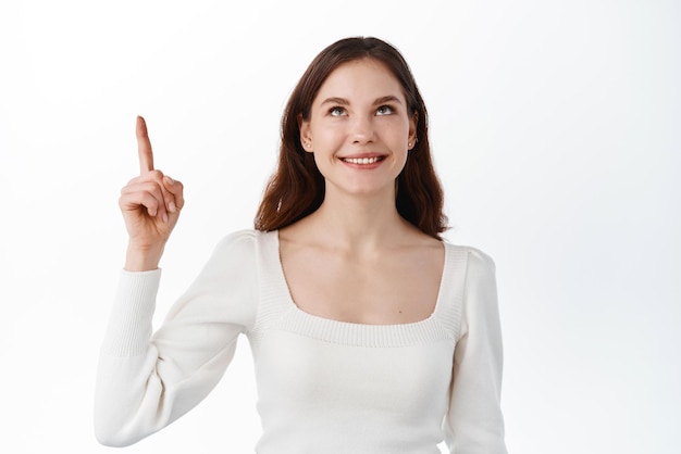 Modelo de linda garota caucasiana apontando os dedos para cima olhando para o anúncio superior com um sorriso satisfeito sonhador mostrando um bom negócio promocional em pé sobre fundo branco