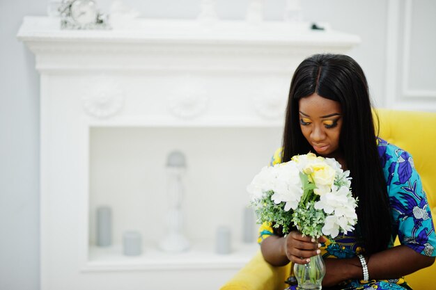 Modelo de mulher afro-americana de elegância veste roupas coloridas Mulheres afro chiques na sala sentada na cadeira amarela com flores na mão