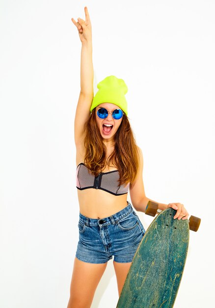 Modelo de mulher elegante sorridente jovem em roupas de moda praia casual e gorro amarelo posando com mesa de longboard, mostrando sinal de rock and roll. Isolado no branco