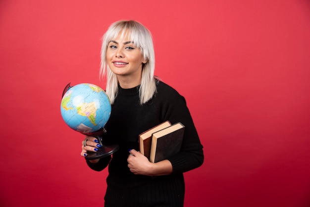 Modelo de mulher jovem posando com livros e um globo terrestre em uma parede vermelha.