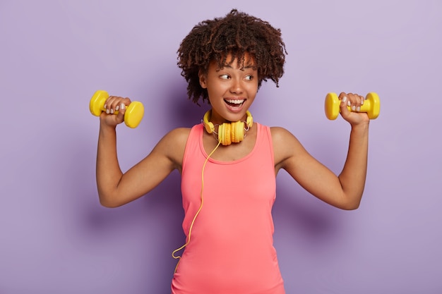 Modelo de pele escura satisfeita com cabelo encaracolado, vestida com uma camisa casual rosa, levanta os braços com halteres, treina músculos, ouve música com fones de ouvido