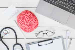 Foto grátis modelo do cérebro rosa; pílulas; estetoscópio; caneta; óculos; prancheta; termômetro; seringa e laptop na mesa de madeira