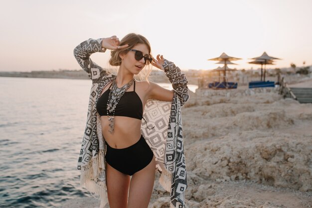 Modelo fascinante posando na praia, pôr do sol. Garota sexy vestindo biquíni preto, maiô com cintura alta, casaquinho, capa com enfeites, linda praia, mar, pedra.