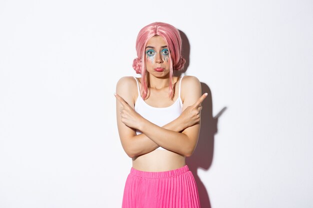 Modelo feminina boba indecisa de peruca rosa, com maquiagem colorida para festa de halloween, apontando para o lado