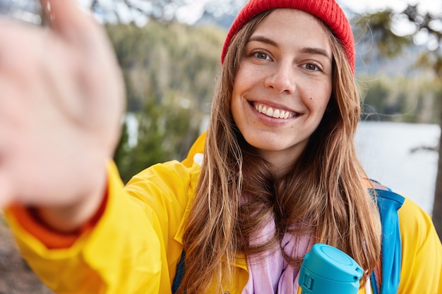 Modelo feminina otimista com um sorriso cheio de dentes, esticando a mão enquanto faz uma selfie, vestida casualmente