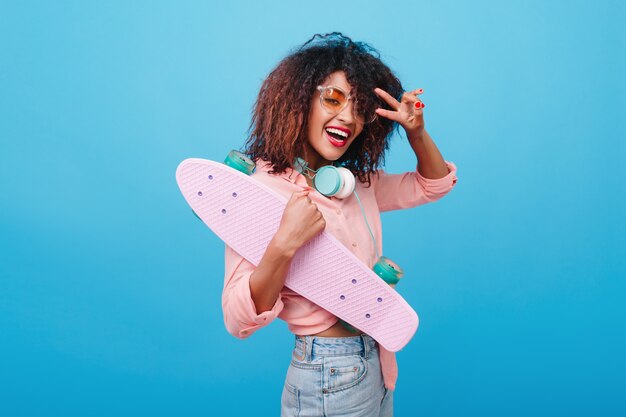 Modelo feminino africano alegre em jeans posando com o símbolo da paz segurando longboard. Mulata rindo com blusa de algodão e fones de ouvido