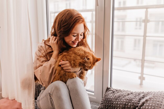 Modelo feminino agradável segurando o gato vermelho de joelhos. Retrato interior da encantadora mulher gengibre posando com o animal de estimação.