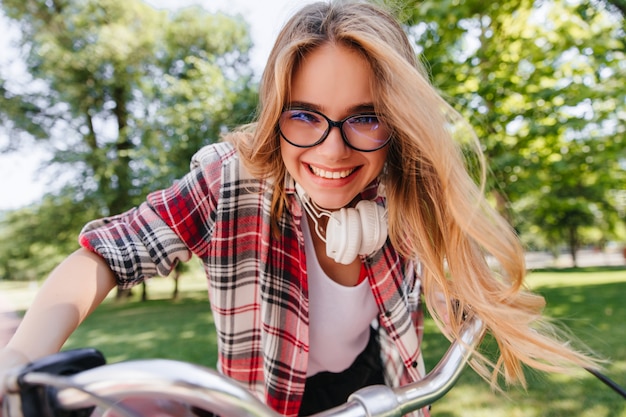 Modelo feminino animado de óculos e fones de ouvido, andando pelo parque. menina loira emocional sentada na bicicleta e rindo.
