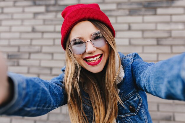 Modelo feminino animado usa chapéu vermelho, fazendo selfie na parede de tijolos. rindo menina branca em óculos escuros e jaqueta jeans posando perto da parede.