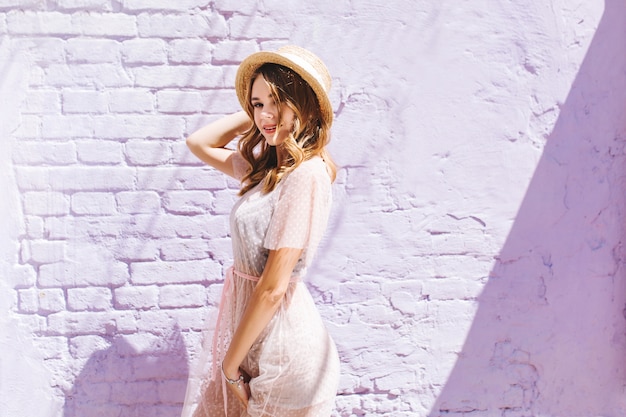 Foto grátis modelo feminino bonito e magro em um vestido estiloso posando em frente a uma parede de tijolos e sorrindo com uma expressão facial astuta