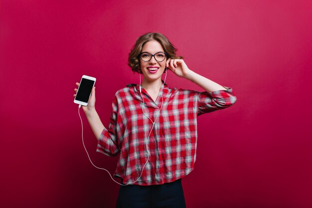 Modelo feminino branco entusiasmado com penteado curto da moda, posando com telefone Menina elegante em camisa xadrez clarete segurando o smartphone.