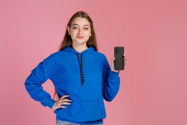 Modelo feminino confiante, mantendo uma mão na cintura e demonstrando smartphone moderno na câmera