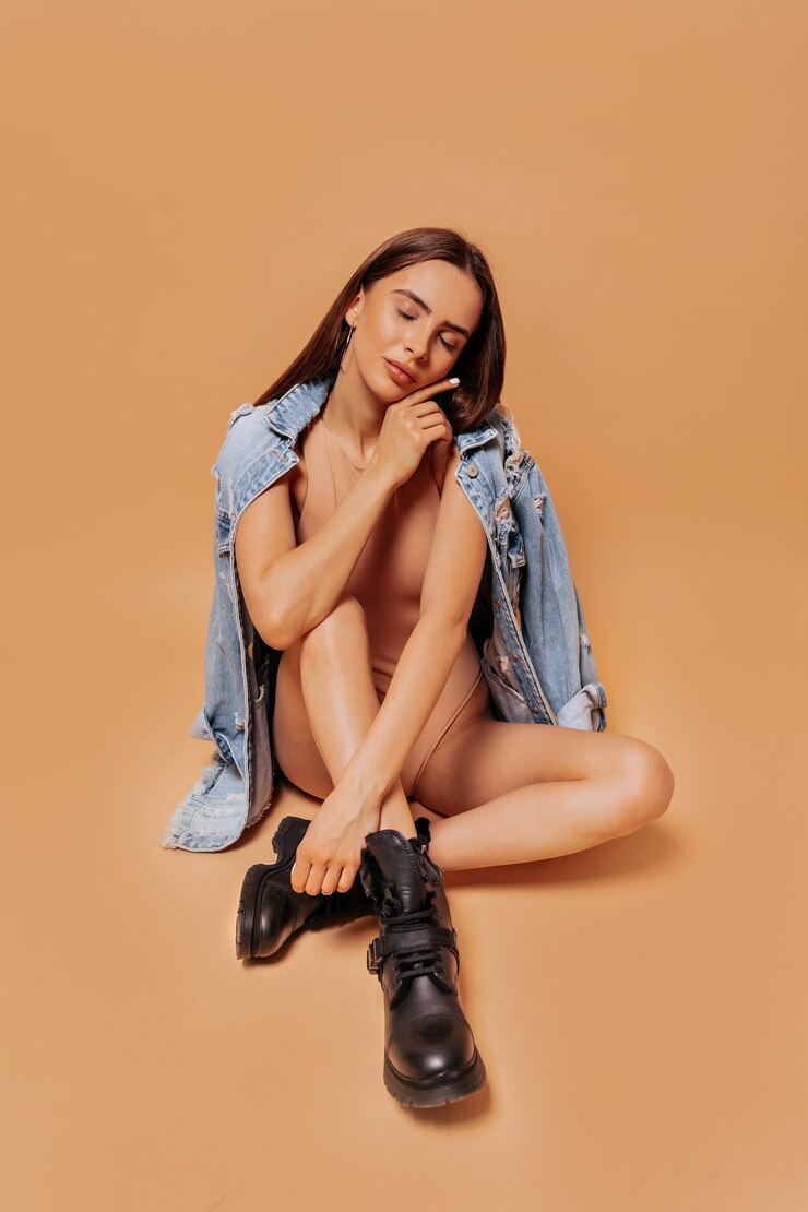Foto grátis modelo feminino elegante vestindo jaqueta jeans e botas, sentado na parede bege com um sorriso adorável e olhos fechados