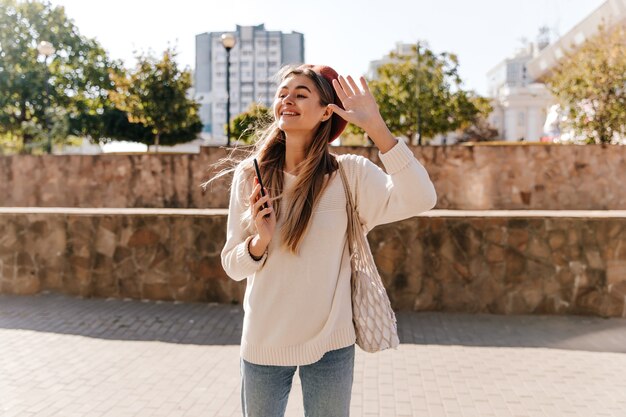 Modelo feminino emocional na boina, acenando com a mão no fundo da cidade. Senhora bem vestida alegre, relaxando ao ar livre no dia de outono.