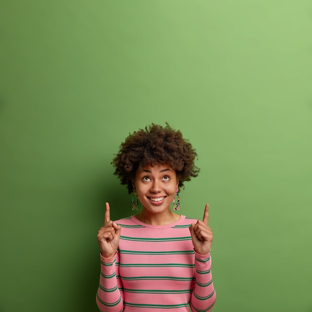 Modelo feminino étnico positivo atrai sua atenção para cima, aponta os dedos indicadores para o espaço vazio, sugere clicar ou se inscrever, mostra espaço para promoção