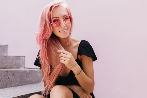 Modelo feminino europeu sensual tocando seu cabelo rosa enquanto relaxa nas escadas.