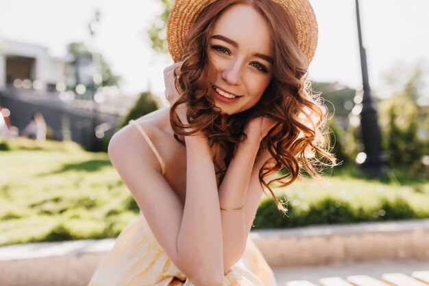 Foto grátis modelo feminino glamoroso com cabelo ruivo, posando com um sorriso fofo na natureza. foto ao ar livre de menina bem-humorada com chapéu de palha, apreciando a sessão de fotos no parque.