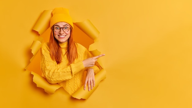 Modelo feminino jovem alegre tem um sorriso dentuço de cabelo vermelho apontando para o espaço da cópia, vestido com óculos de camisola de suéter amarelo.