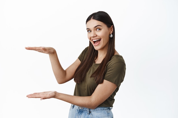 Modelo feminino morena alegre segurando um espaço de cópia vazio para seu logotipo ou anúncio de produto, fazendo um gesto de caixa grande, parede branca