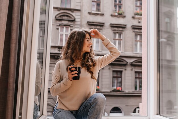 Modelo feminino muito sorridente de óculos sentado no parapeito com uma xícara de café