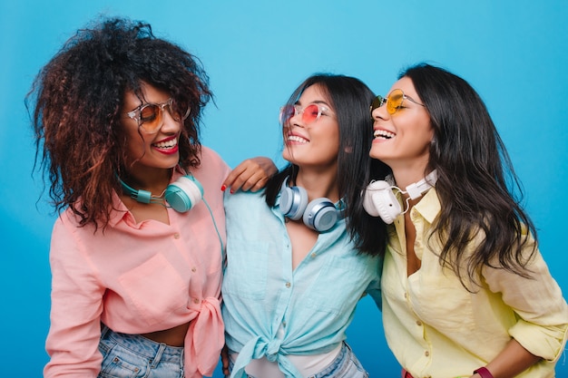 Foto grátis modelo feminino mulato interessado olhando com sorriso para meninas asiáticas usa fones de ouvido grandes. mulheres na moda em roupas coloridas conversando.