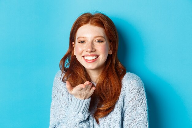 Modelo feminino ruiva adorável sorrindo, mandando beijo no ar para a câmera, em pé contra um fundo azul.