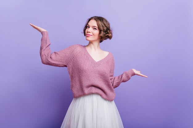 Modelo feminino satisfeito com a expressão de rosto feliz, posando com roupas de inverno e sorrindo. Mulher de cabelo curto no lenço que expressa emoções positivas na parede roxa.