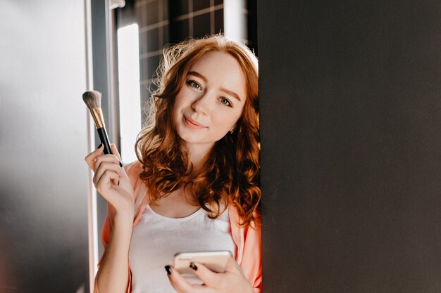 Modelo feminino sensual com pincel de maquiagem, posando em casa. Menina ruiva encaracolada segurando o telefone e olhando.