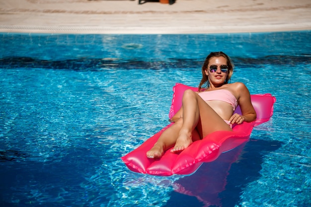 Modelo feminino sexy, descansando e tomando banho de sol em um colchão na piscina. mulher em um maiô de biquíni rosa flutuando sobre um colchão inflável rosa. fps e protetor solar