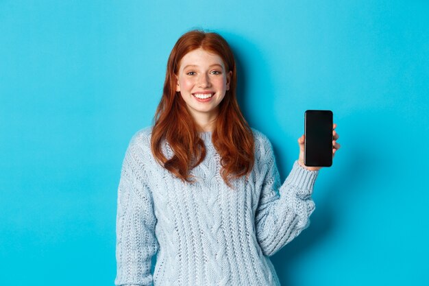 Modelo feminino sorridente com cabelo vermelho mostrando a tela do smartphone, segurando o telefone e demonstrando o aplicativo, em pé sobre um fundo azul