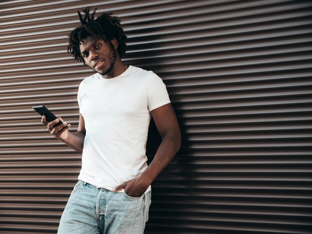 Modelo hipster bonito Homem africano não barbeado vestido com camiseta branca de verão Homem fashion com penteado dreadlocks Posando na ruaOlhando para a tela do smartphone usando aplicativos de celular