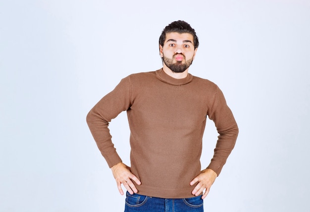 modelo jovem bonito em pé com uma camisola marrom e posando com as mãos nos quadris, sobre uma parede branca.