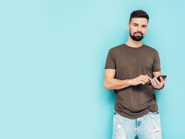 Modelo lambersexual bonito e elegante hipster homem vestido de camiseta e jeans Moda masculina isolada na parede azul no estúdio Segurando smartphone olhando para a tela do celular usando aplicativos