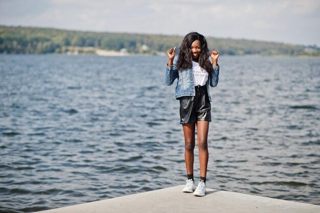 Modelo magro de pele escura afro-americana posou em um short preto e jaqueta jeans contra o lado do mar