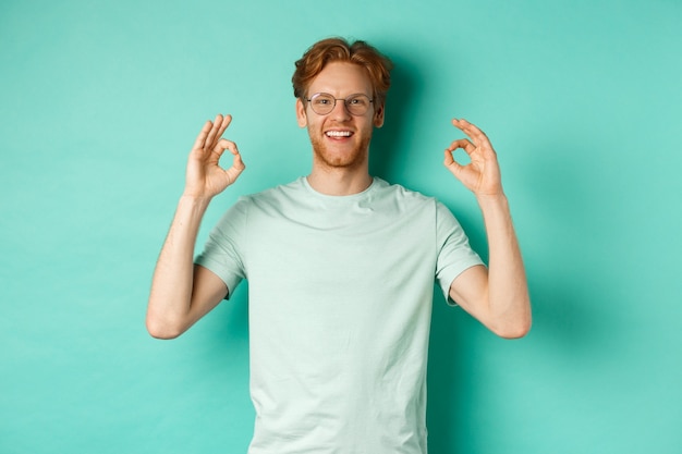 Modelo masculino atraente, com cabelo vermelho, usando óculos, mostrando o sinal de ok em aprovação e dizendo que sim, sorrindo satisfeito, em pé sobre o fundo da casa da moeda.