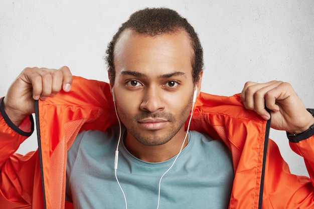 Modelo masculino atraente e elegante com cerdas, ouve música, usa fones de ouvido, mantém as mãos no anoraque laranja,