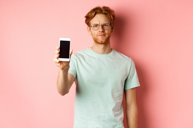 Modelo masculino desapontado com a testa franzida, mostrando a tela do smartphone, em pé sobre um fundo rosa