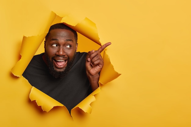 Foto grátis modelo masculino preto alegre e feliz aponta o dedo indicador para o lado, mantém a boca aberta, mostra a língua, atrai sua atenção para o espaço em branco