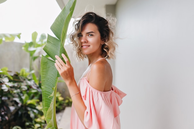 Modelo muito feminino com olhos azuis, olhando e segurando uma folha verde. adorável garota bronzeada com expressão de rosto calmo em pé no quintal.