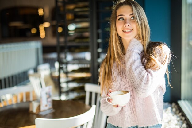 Modelo muito jovem estudante adolescente vestida com jeans de roupas casuais no café segurando uma xícara de chá de café nas mãos