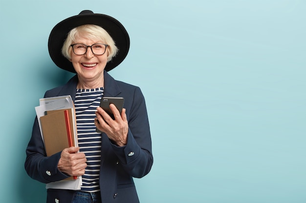 Moderadora feminina alegre socializa online, segura um gadget moderno, recebe notificações, segura papéis com um bloco de notas, vestida com roupa formal elegante, isolada sobre uma parede azul, espaço em branco