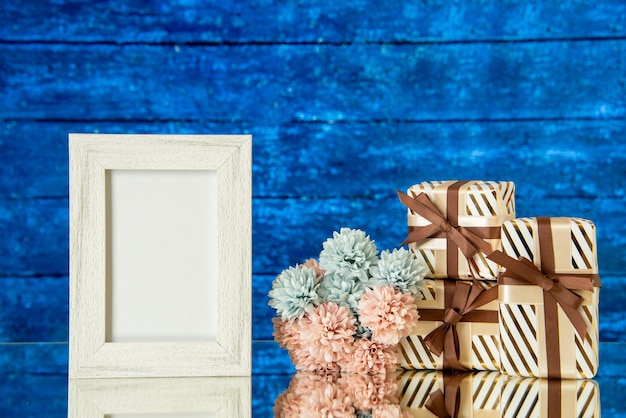 Moldura de foto branca de vista frontal, caixas de presente de férias, flores refletidas no espelho com um fundo azul de madeira