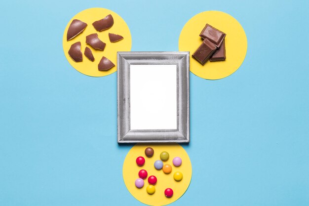 Moldura de prata branca sobre o quadro circular amarelo com doces de gema; pedaços de chocolate e cascas de ovos de Páscoa em pano de fundo azul