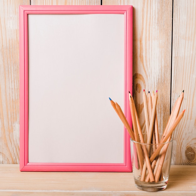 Foto grátis moldura em branco branca com borda rosa e lápis de cor no suporte de vidro na mesa de madeira