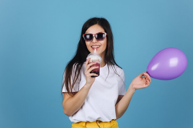 Morena alegre em óculos de sol pretos parece feliz posando com um coquetel e balão